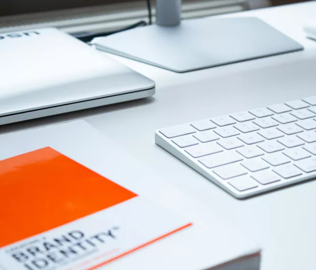 Clean white desk setup with a keyboard, monitor stand, laptop, and a book with an orange accent, suggesting a modern, organized workspace.