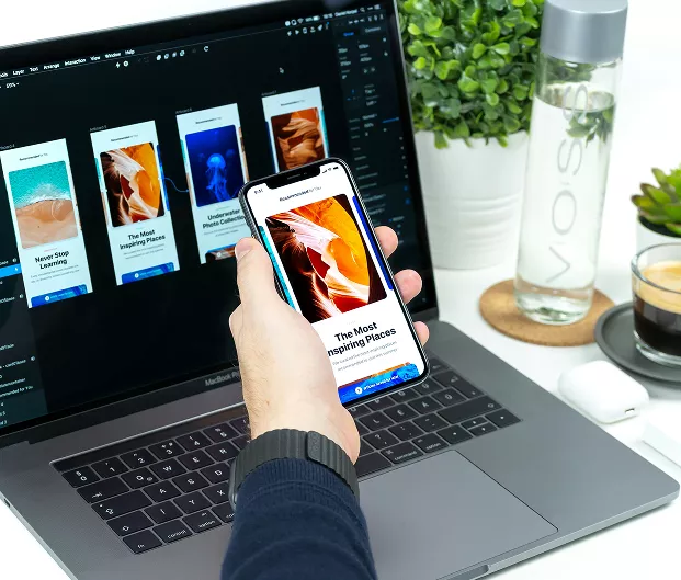 An over-the-shoulder view shows a person holding an iPhone in their left hand, displaying a mobile app design featuring an orange canyon landscape