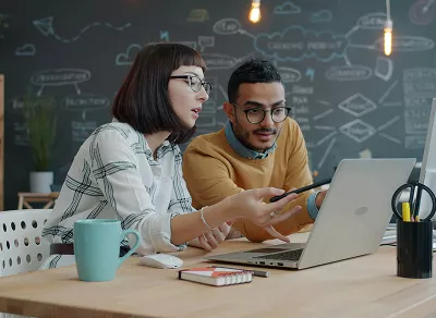 Two coworkers collaborating at a desk, reviewing a laptop screen together in a modern office with notes and sketches on the wall.