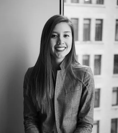 B&W portrait of a smiling woman with long hair, wearing a grey blazer, posed in front of a window with city buildings behind.